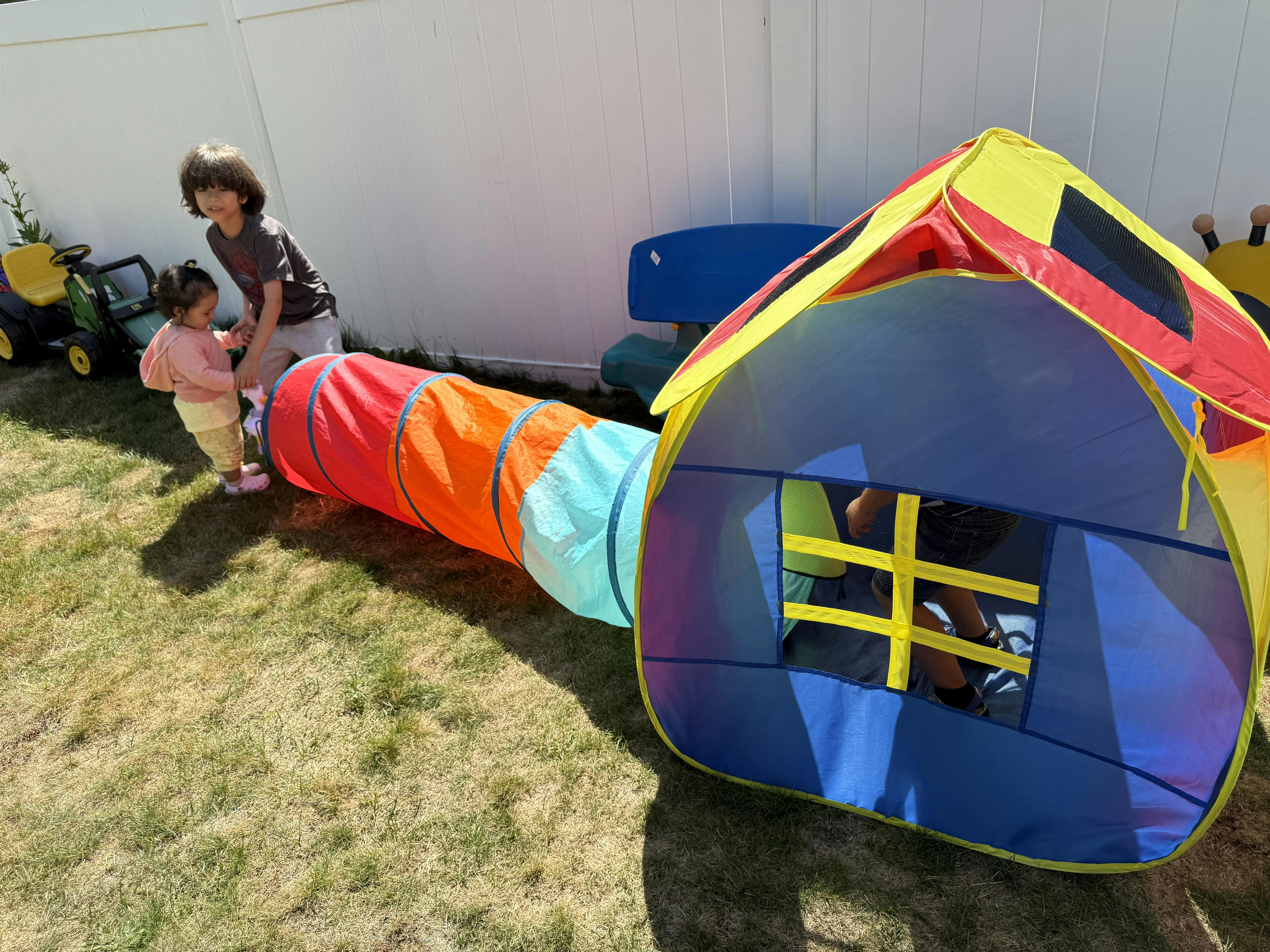 Two children playing with a colorful tunnel and playhouse outdoors