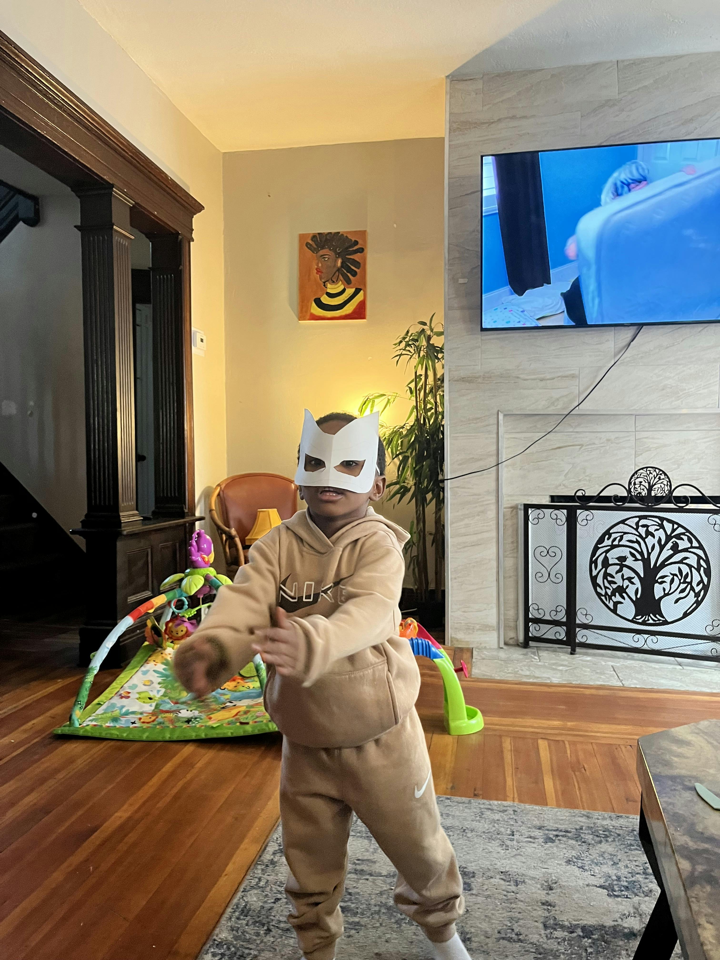 Child wearing a white mask and tan sweatsuit standing indoors