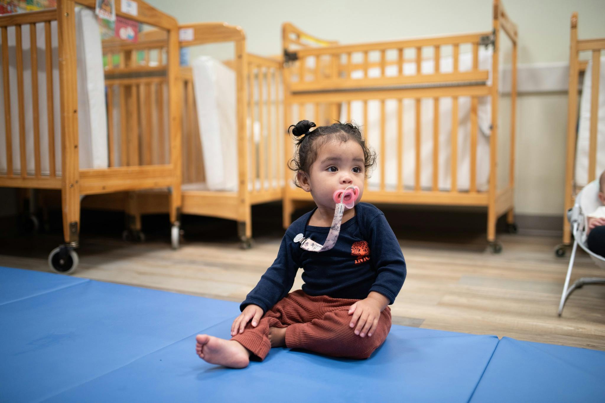 A child with a pacifier sitting on a blue mat in a room with cribs