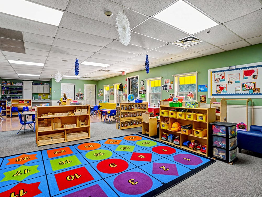 A classroom with light green walls, colorful rug, shelves of toys, and small tables