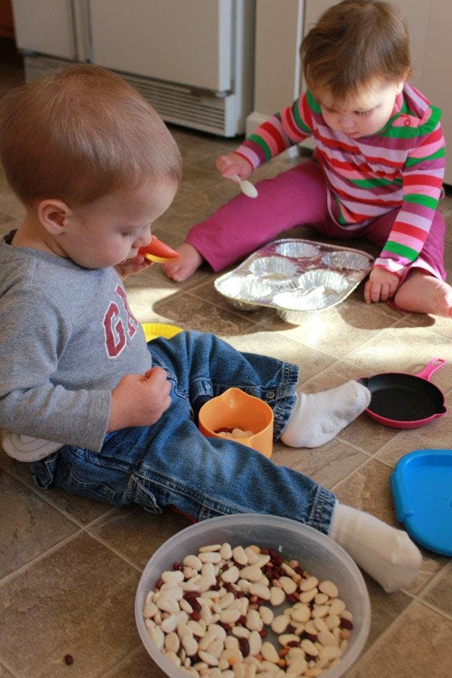 Two children playing on the floor with kitchen items and a bowl of beans