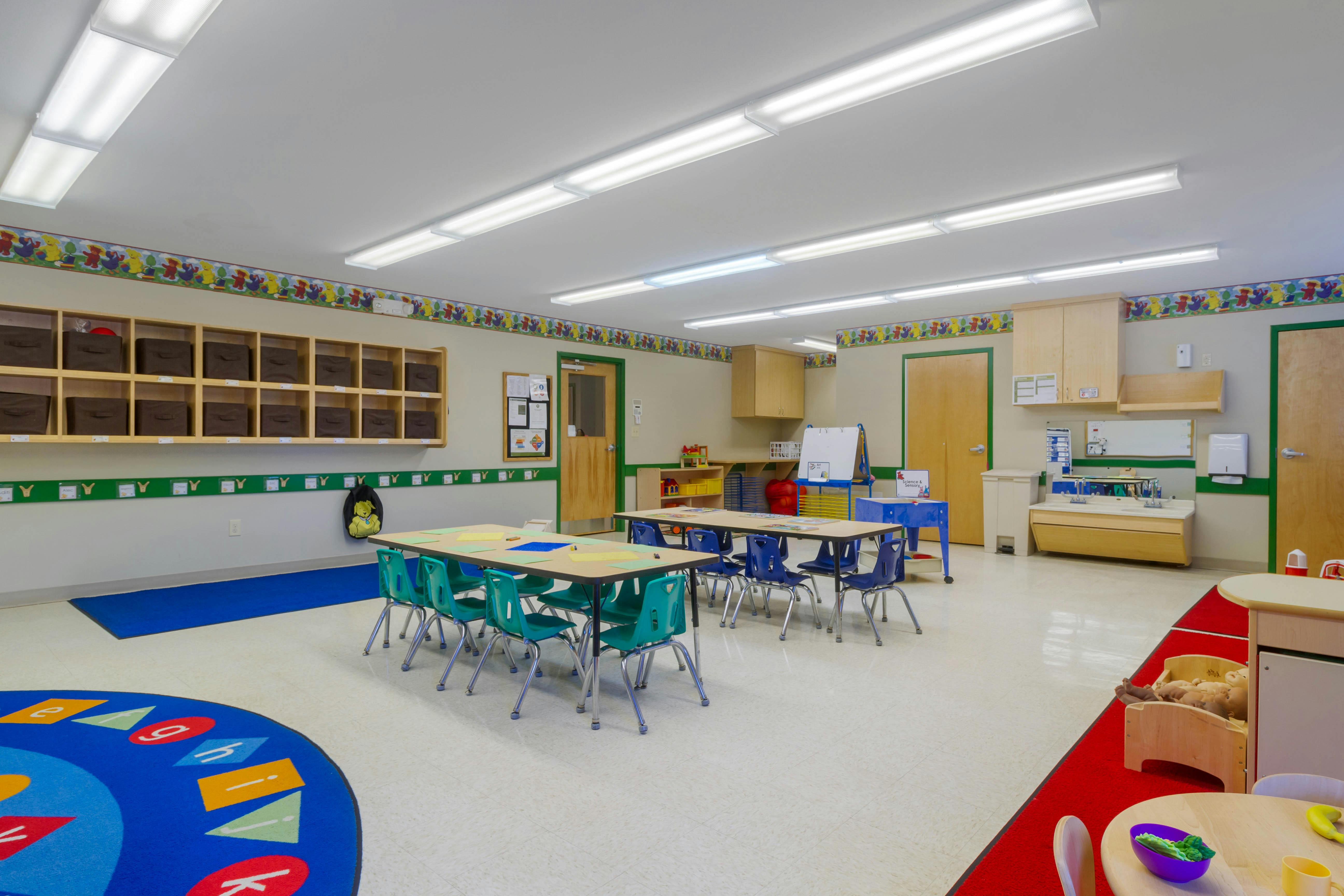 An empty classroom with small tables and chairs, cubbies, and a sink