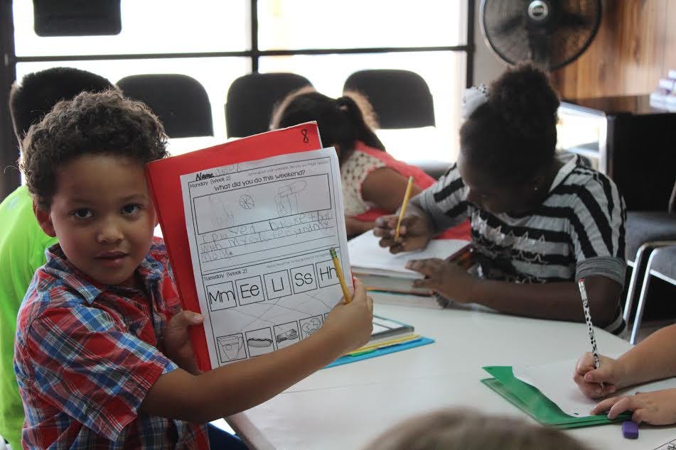 Child holding up a worksheet with other children writing at a table