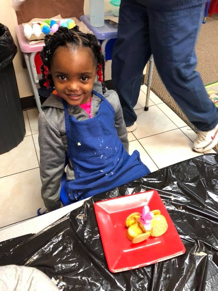 Child in a blue apron smiling, with a red plate of food in front
