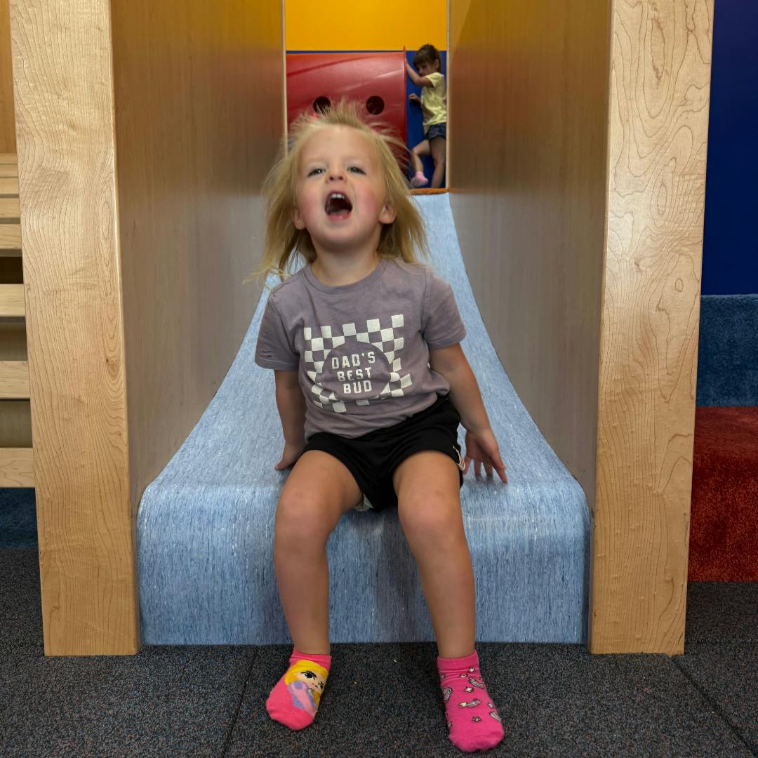 Child sitting on a soft play slide, mouth open, wearing a DAD'S BEST BUD t-shirt