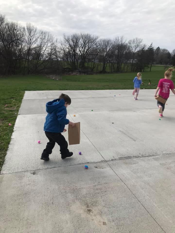 Child in blue jacket holding a bag, collecting colorful eggs on a paved surface