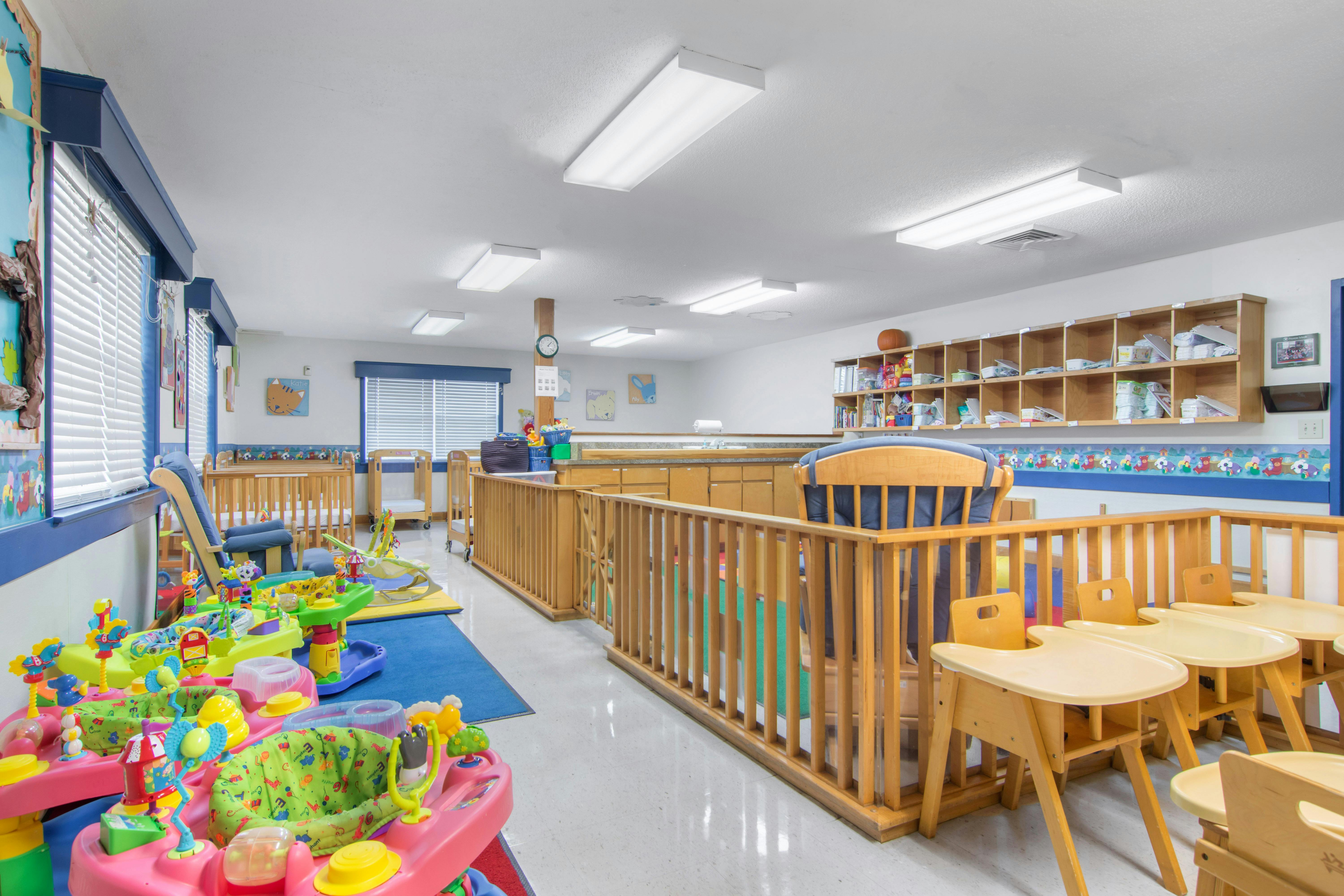 A colorful childcare classroom with cribs, play mats, and small chairs