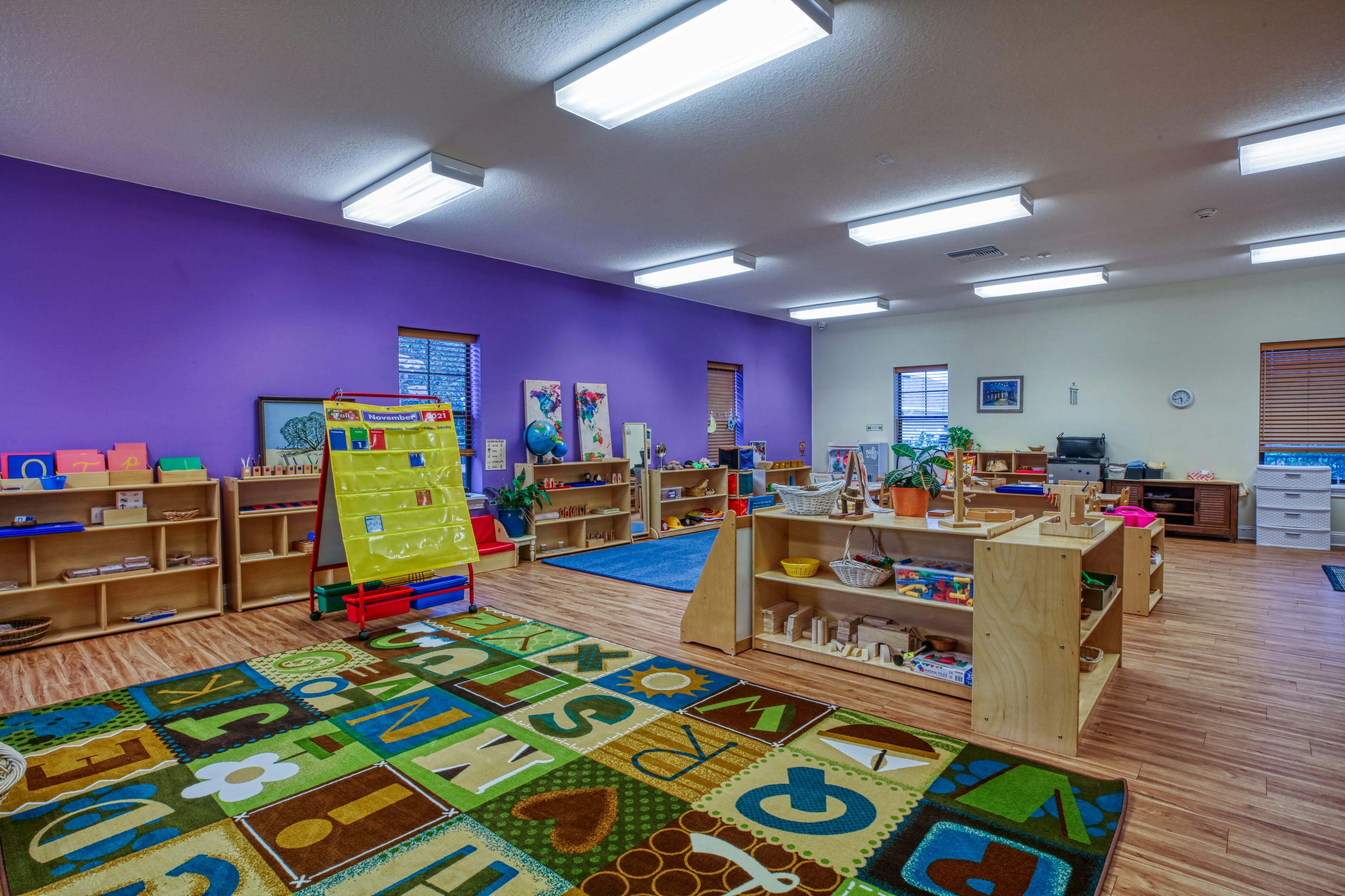 Classroom with purple and off-white walls, wooden shelves, a calendar, and alphabet rug