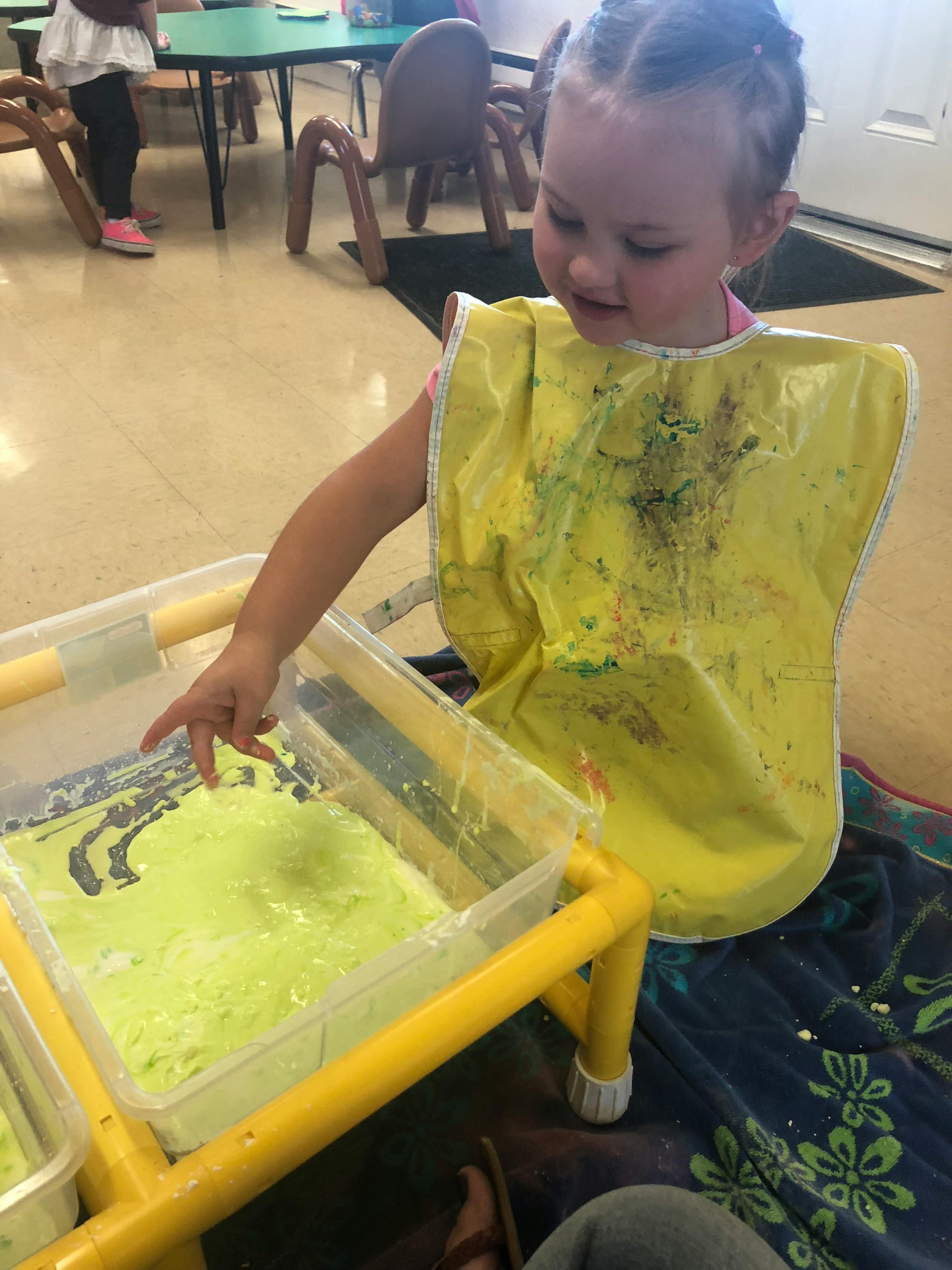 Child in a yellow bib playing with green slime in a clear bin
