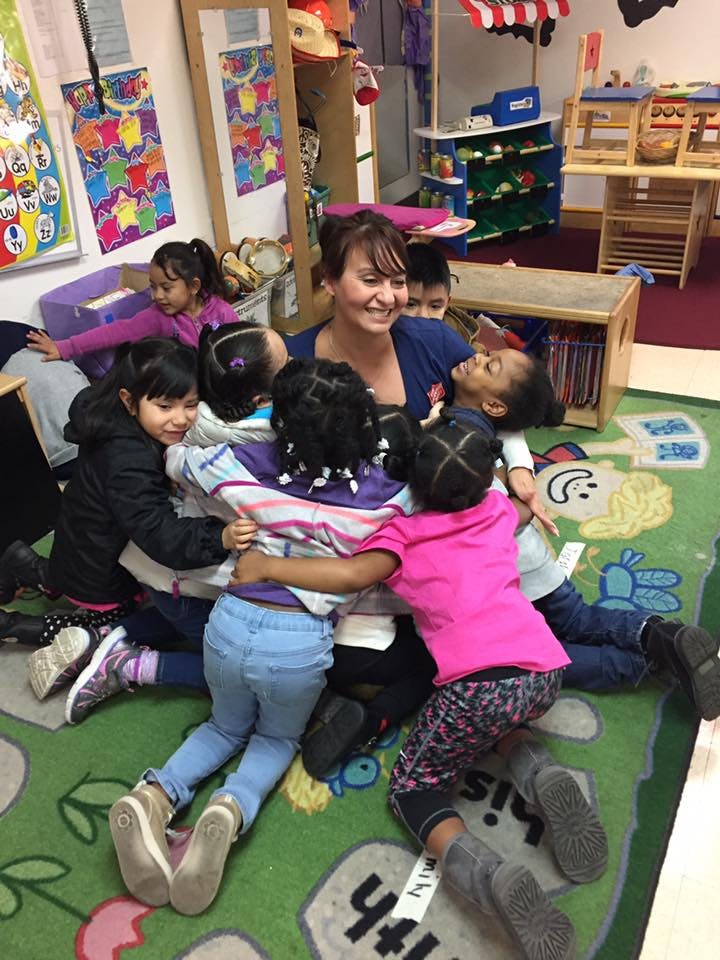 Adult and multiple children in a group hug on a classroom floor