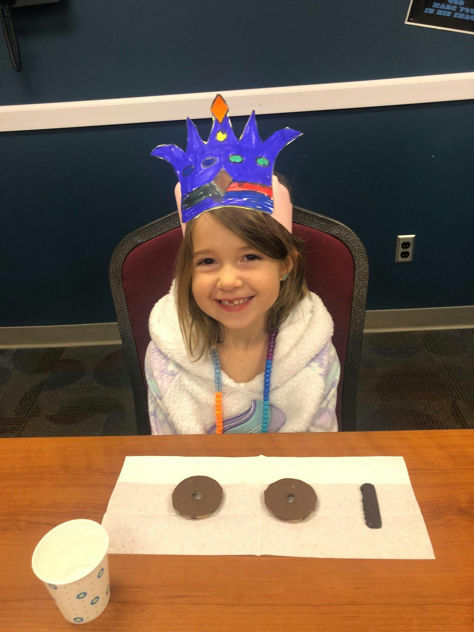 Child wearing a paper crown sitting at a table with a cup and cookies
