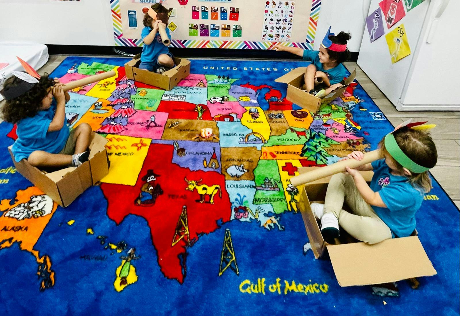 Four children in cardboard boxes on a USA map rug, playing with paper tubes