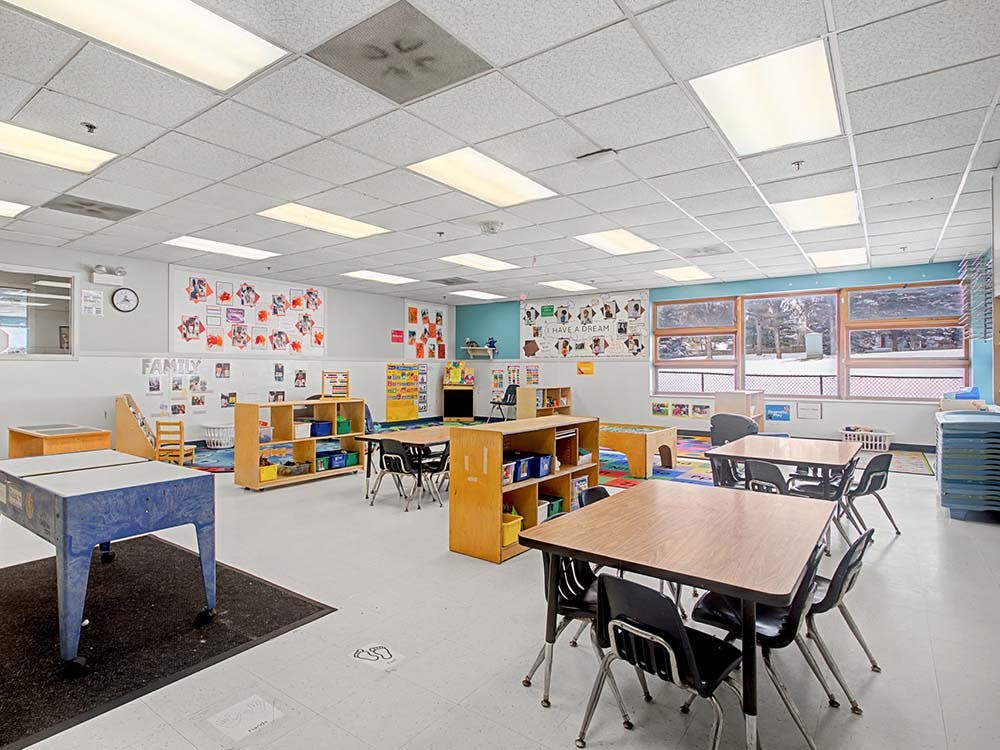 Classroom with tables, chairs, shelving units, and windows showing trees