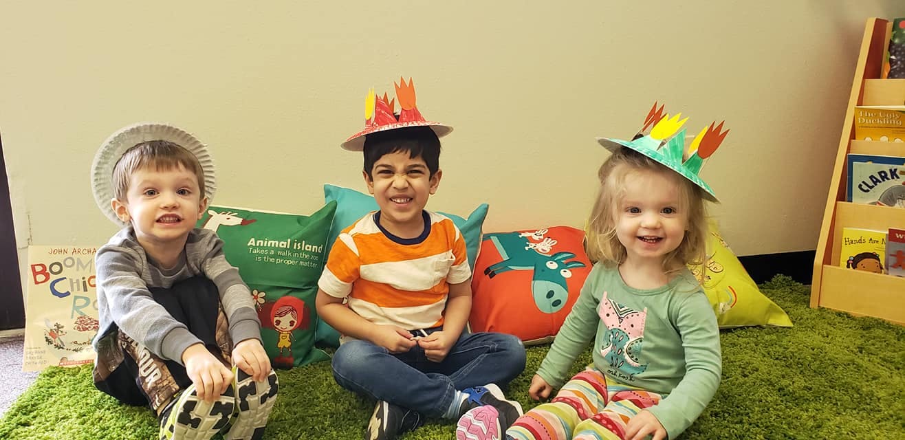 Three children sitting on a green rug, two wearing paper hats