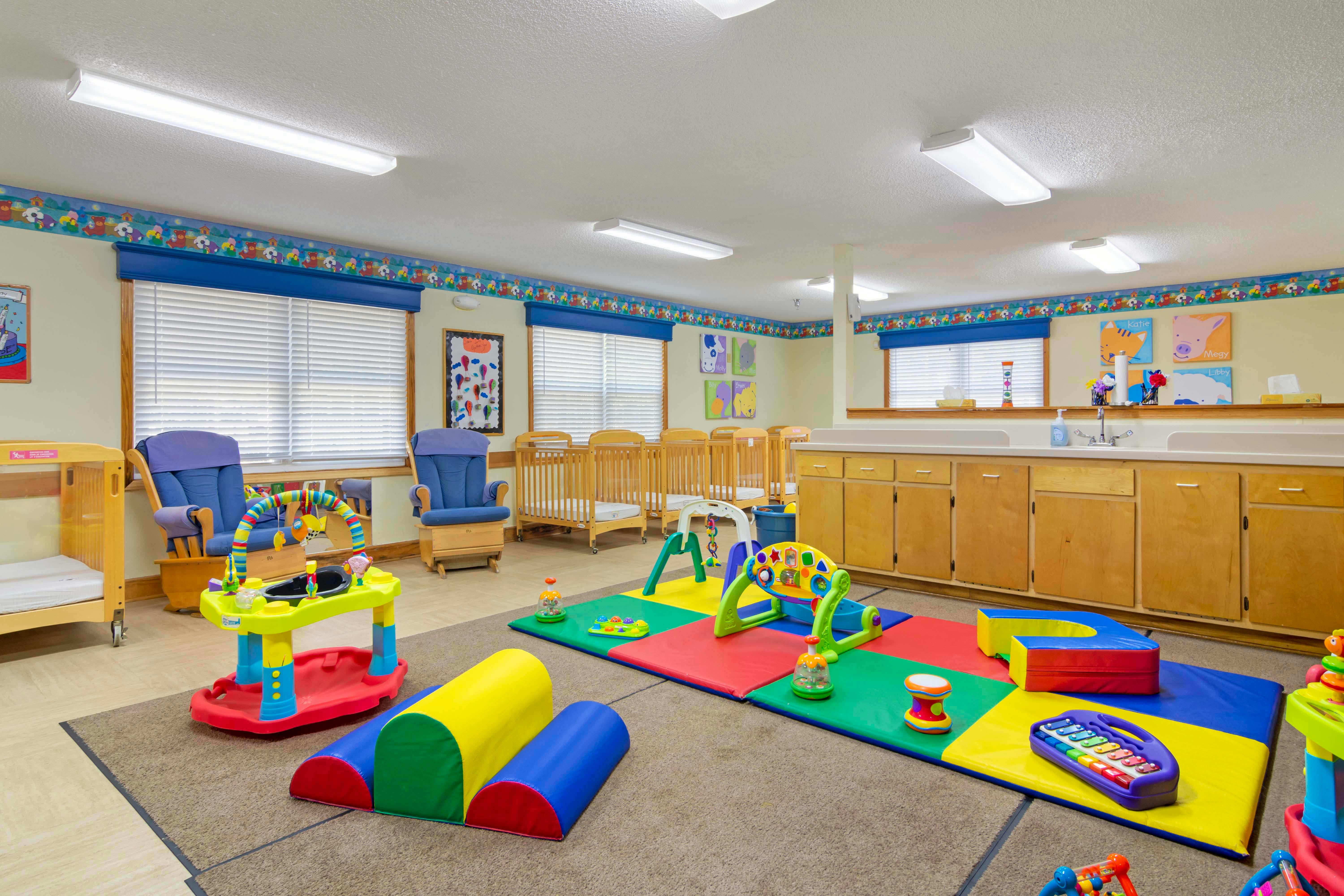 A colorful childcare classroom with cribs, playmats, and toys