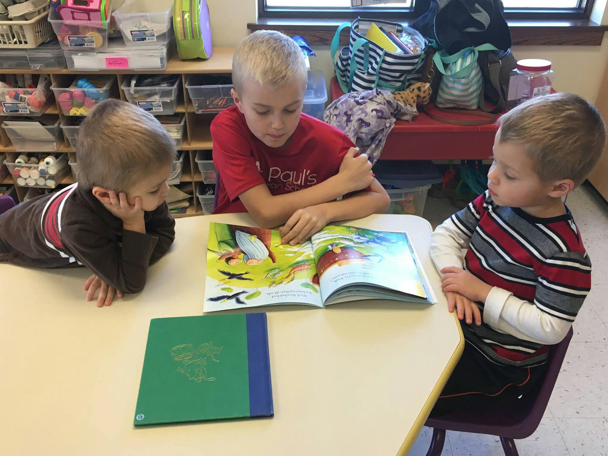 Three children looking at an open book on a table in a classroom