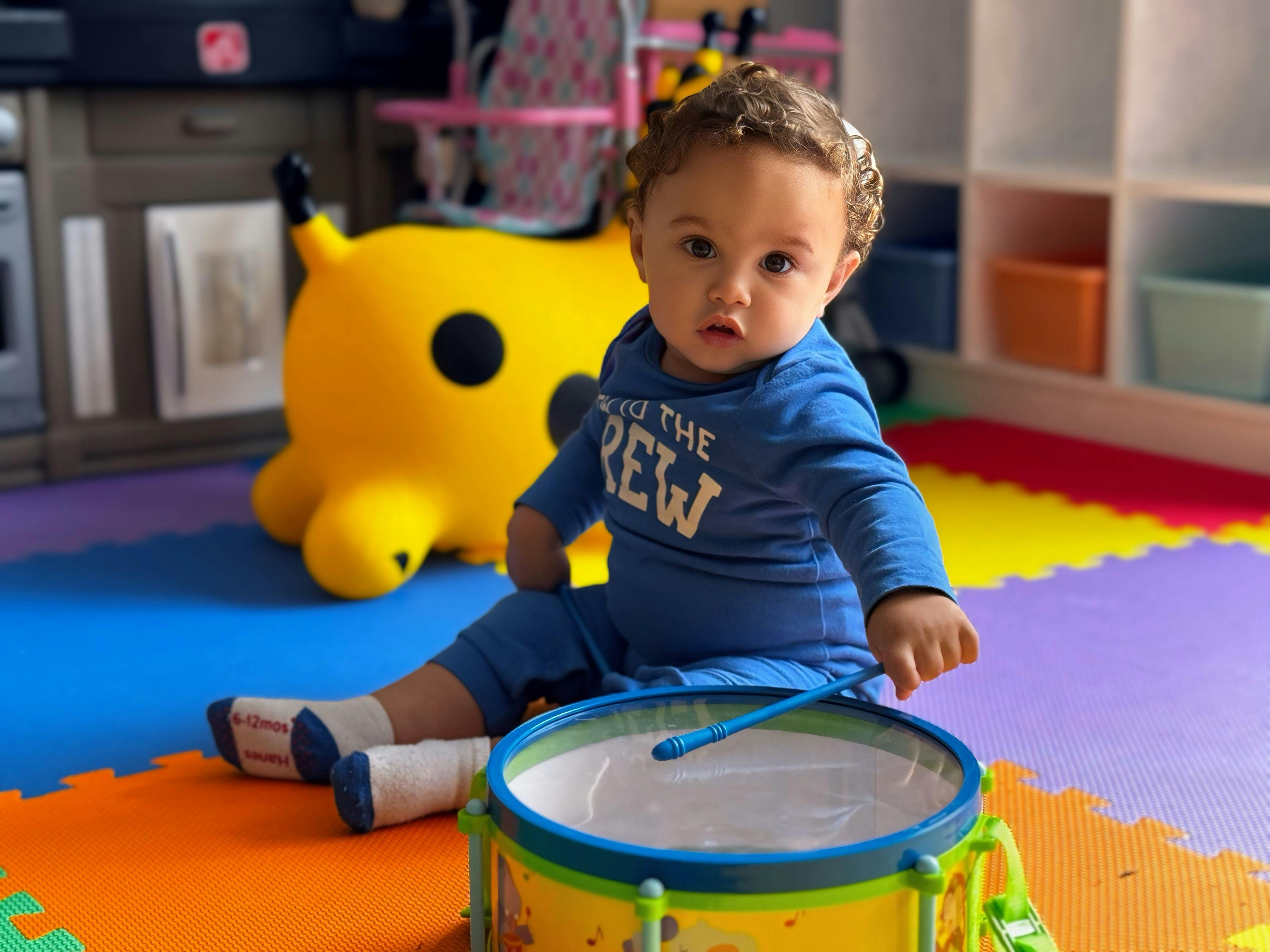 Child sitting on colorful mats, holding a drumstick to a toy drum
