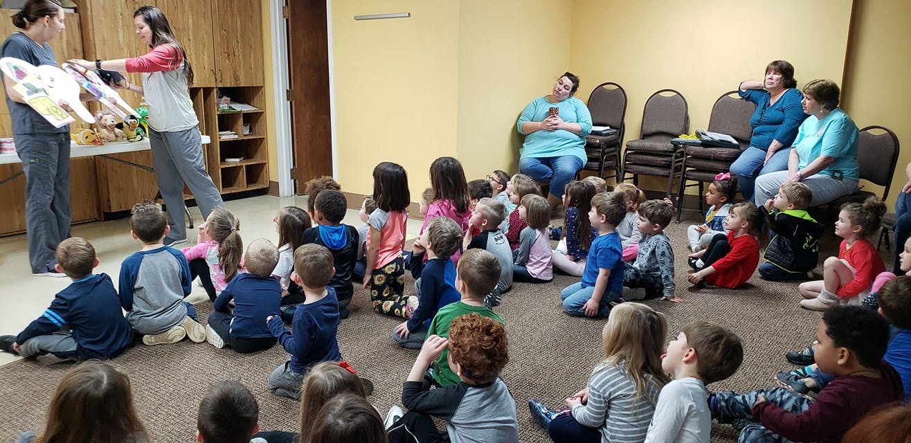 Two people showing a large book to a group of children sitting on the floor