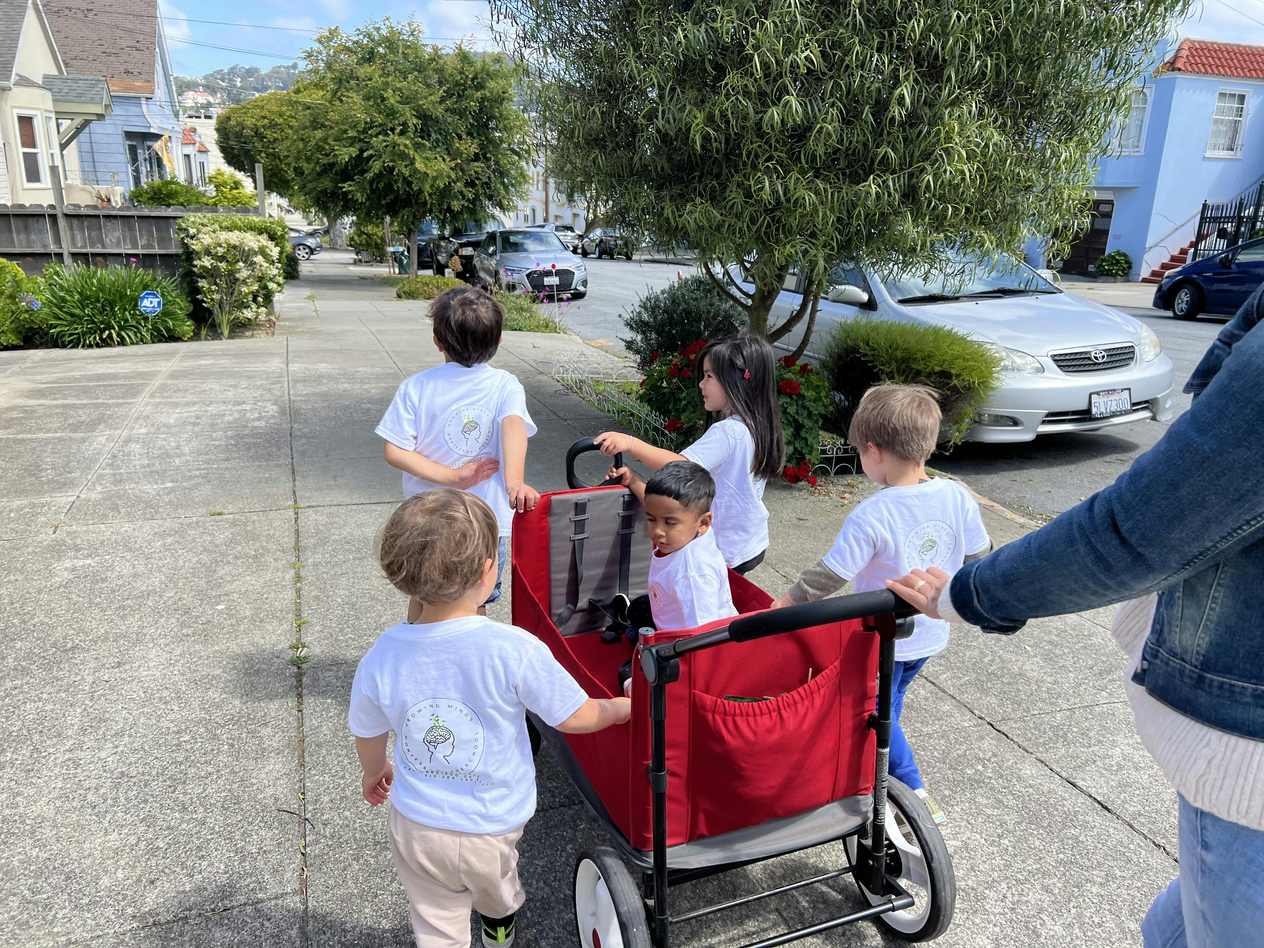 Five children in white t-shirts with an adult pushing a red wagon on a sidewalk