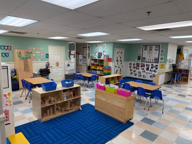 An empty classroom with small tables and chairs, shelves, and a blue rug