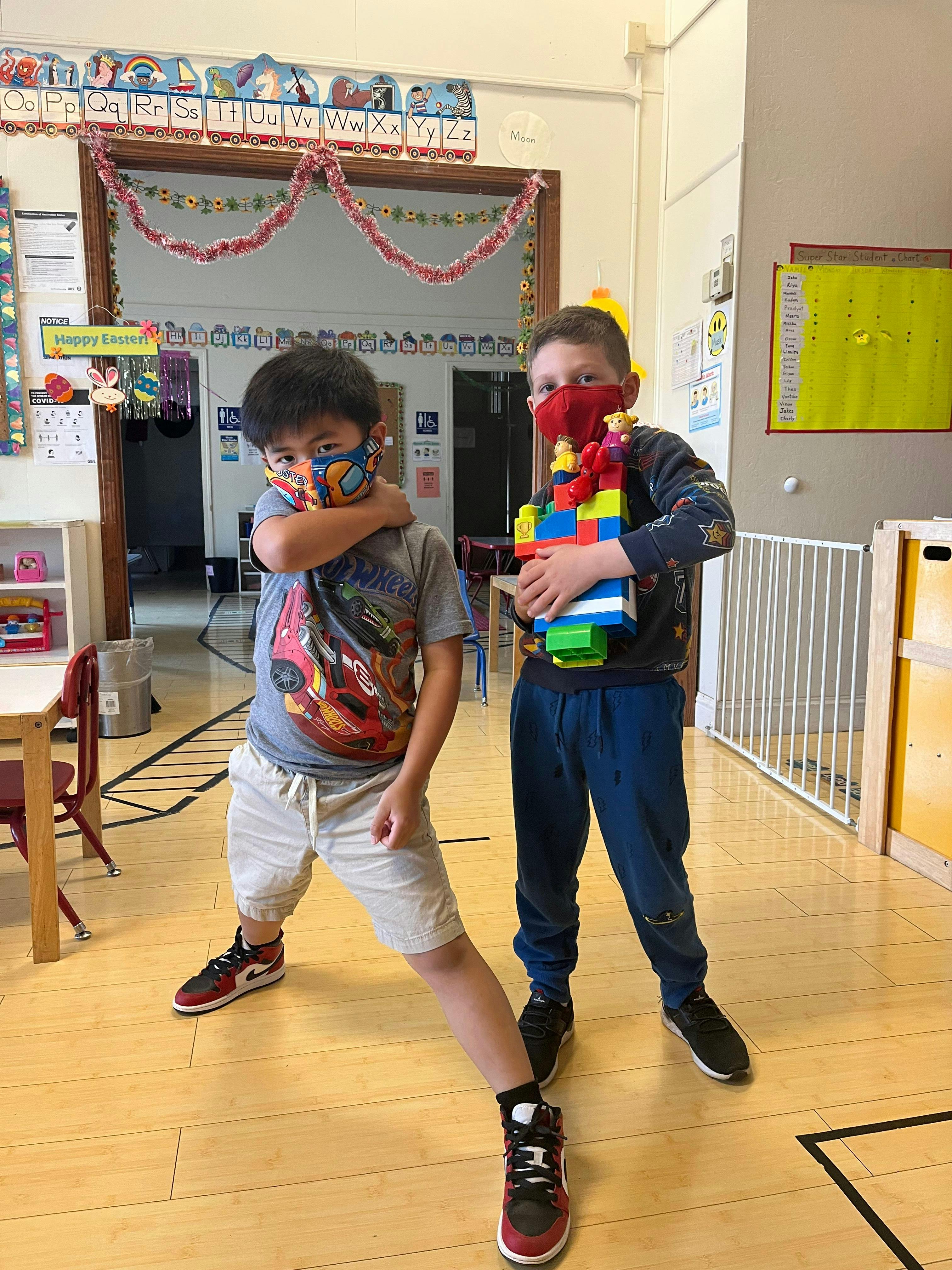 Two children wearing masks posing, one holding a colorful block creation