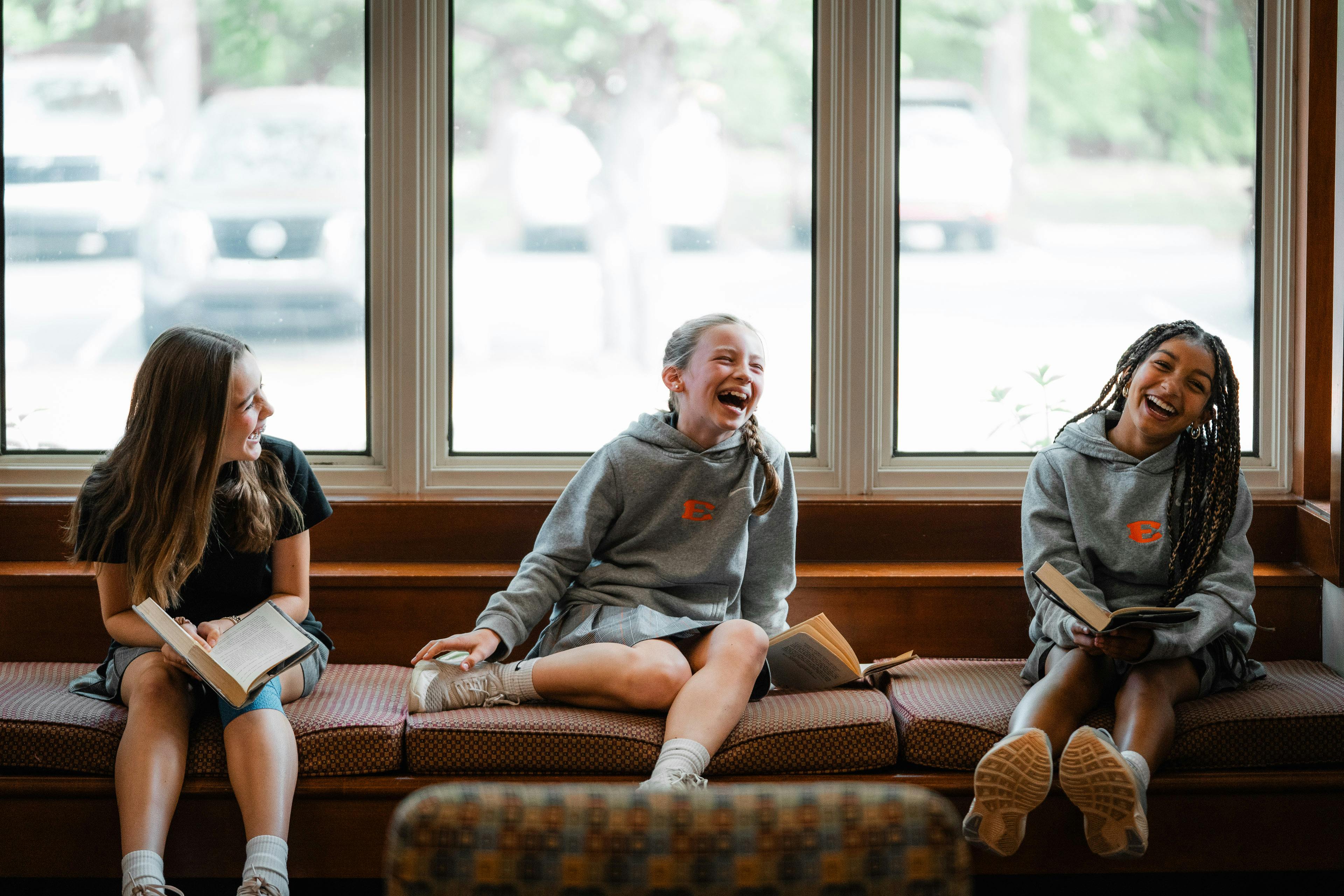 Three teens sitting on a bench by a window reading and laughing