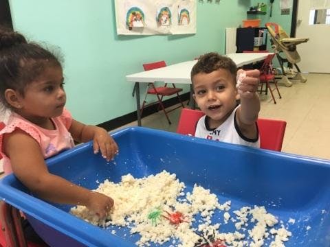 Two children playing with sand in a blue bin
