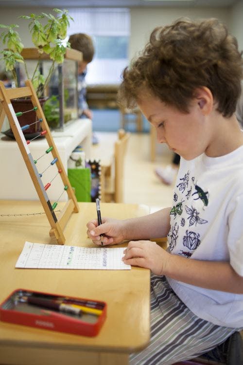 Child writing at a desk with an abacus nearby in a classroom
