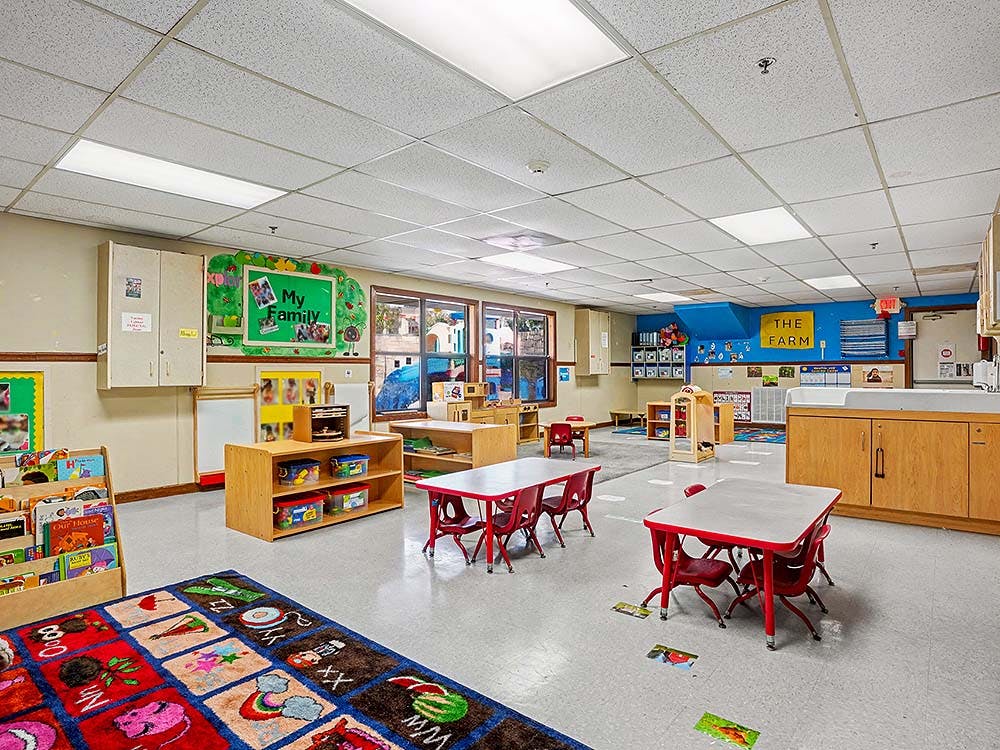 Empty classroom with small tables and chairs, shelves, and learning rug