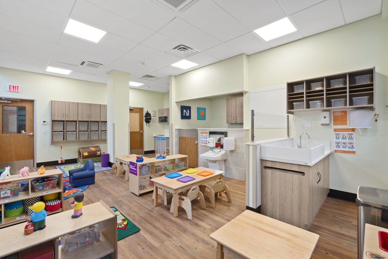 Classroom with small tables, chairs, and shelves of toys, with a changing station