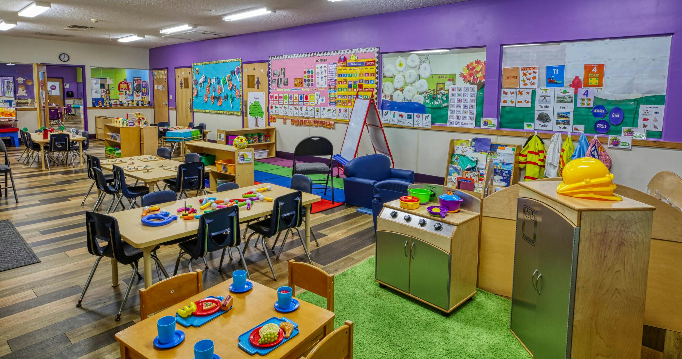 Colorful preschool classroom with several tables and chairs, play kitchen, and educational posters