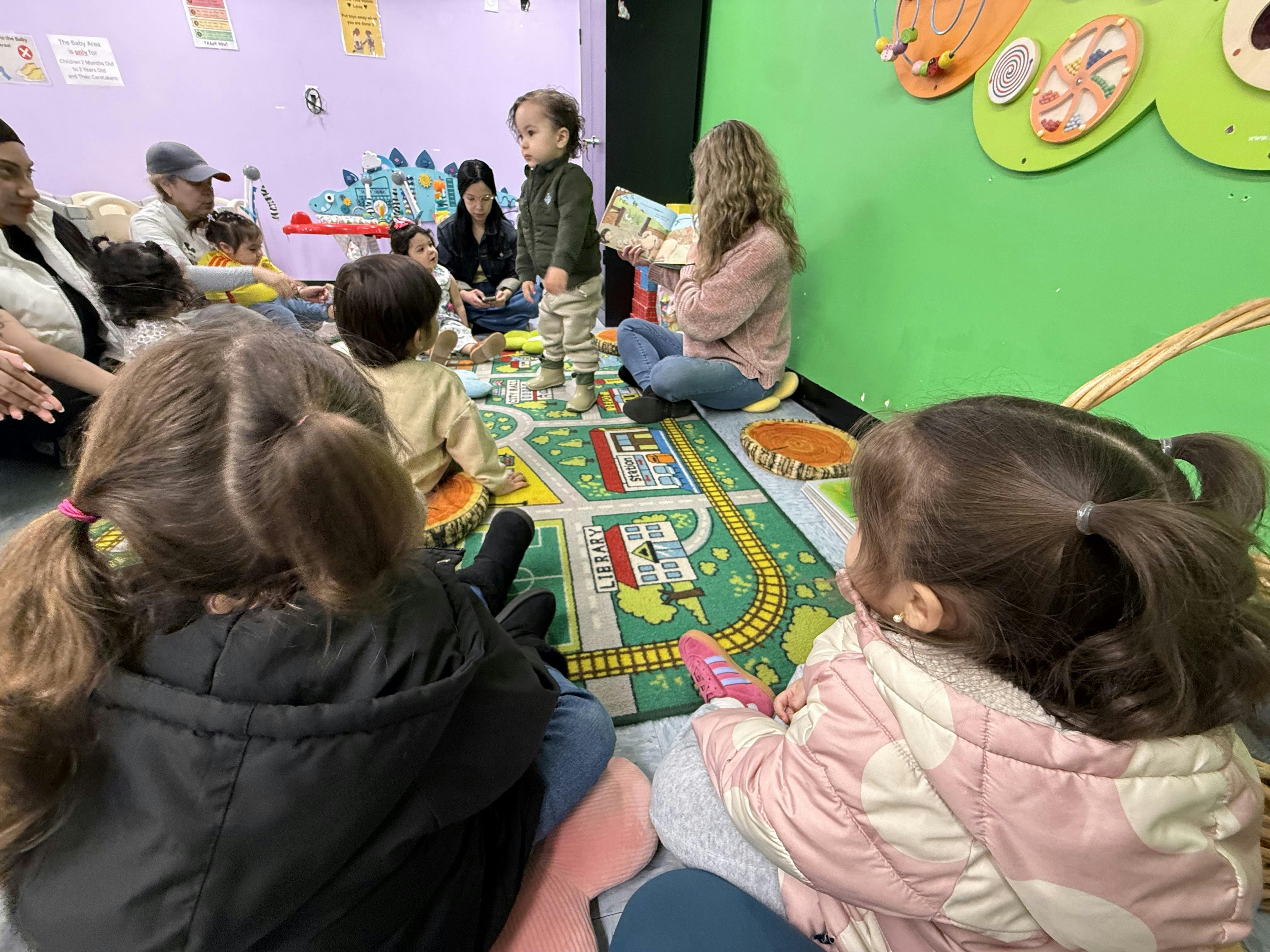 People and children gathered on a rug in a playroom listening to a story