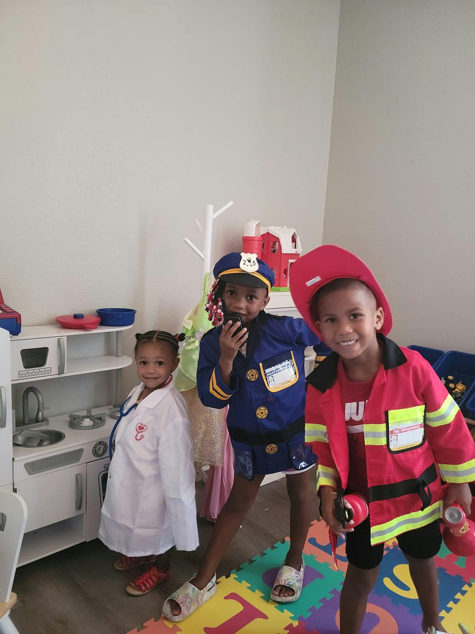 Three children in dress-up costumes, one in a doctor's coat, one in a police uniform, one in a firefighter uniform