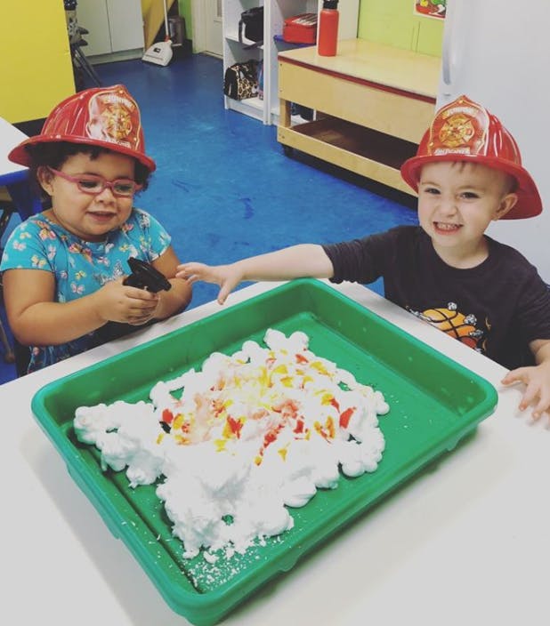 Two children in firefighter hats playing with shaving cream and colored pieces in a tray