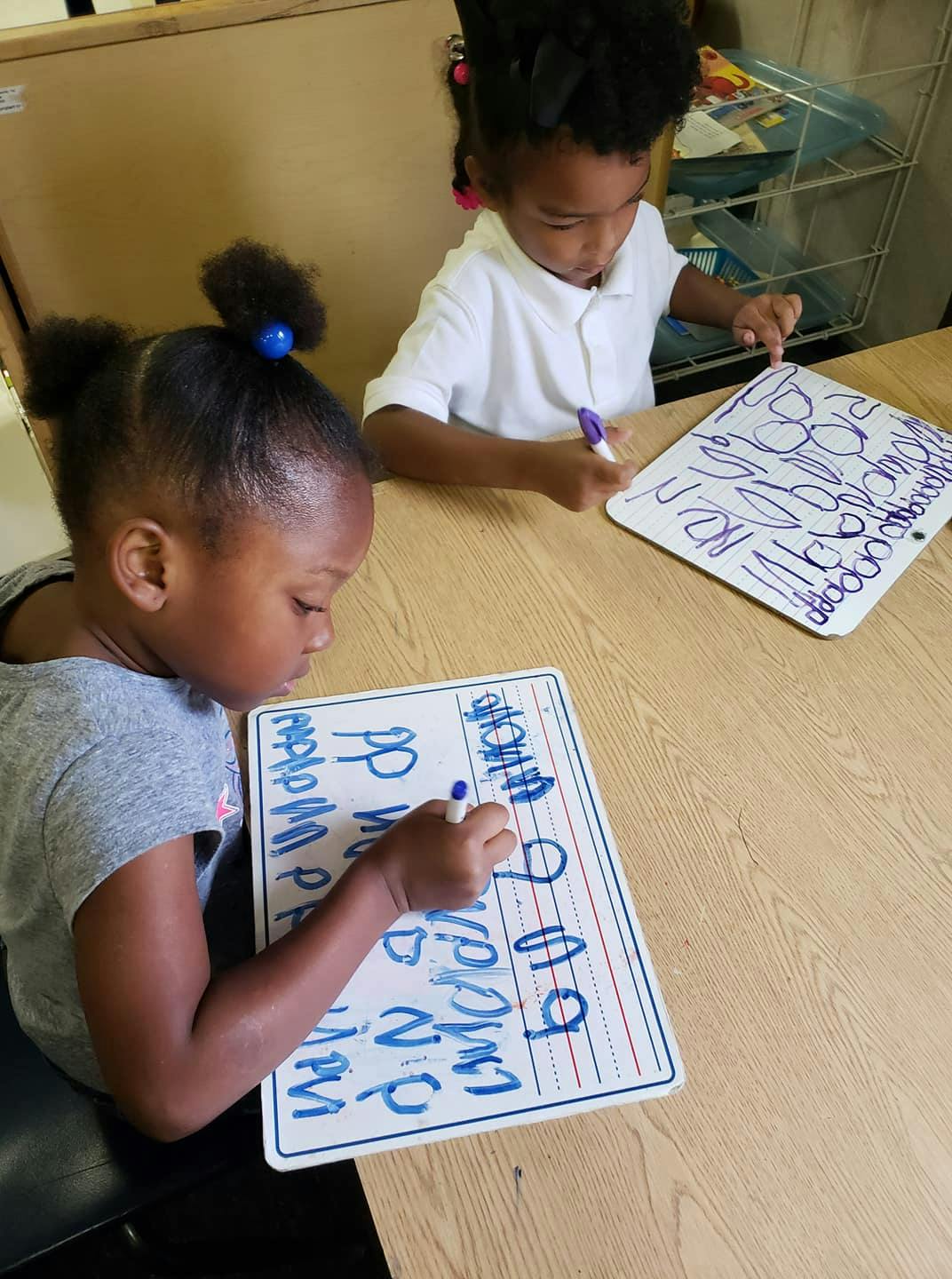 Two children writing on whiteboards at a table