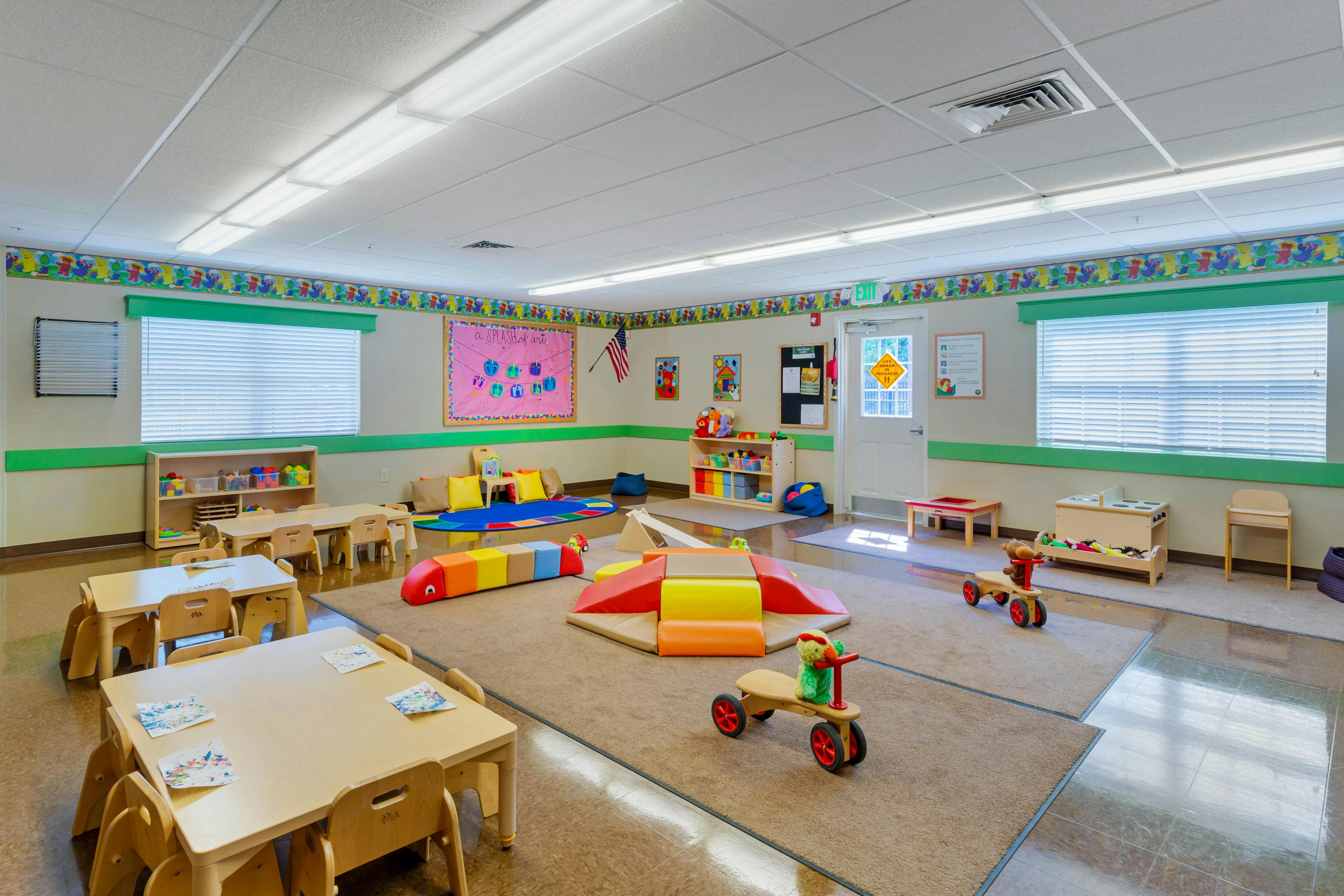 A brightly lit classroom with small tables and chairs, play mats, and toys