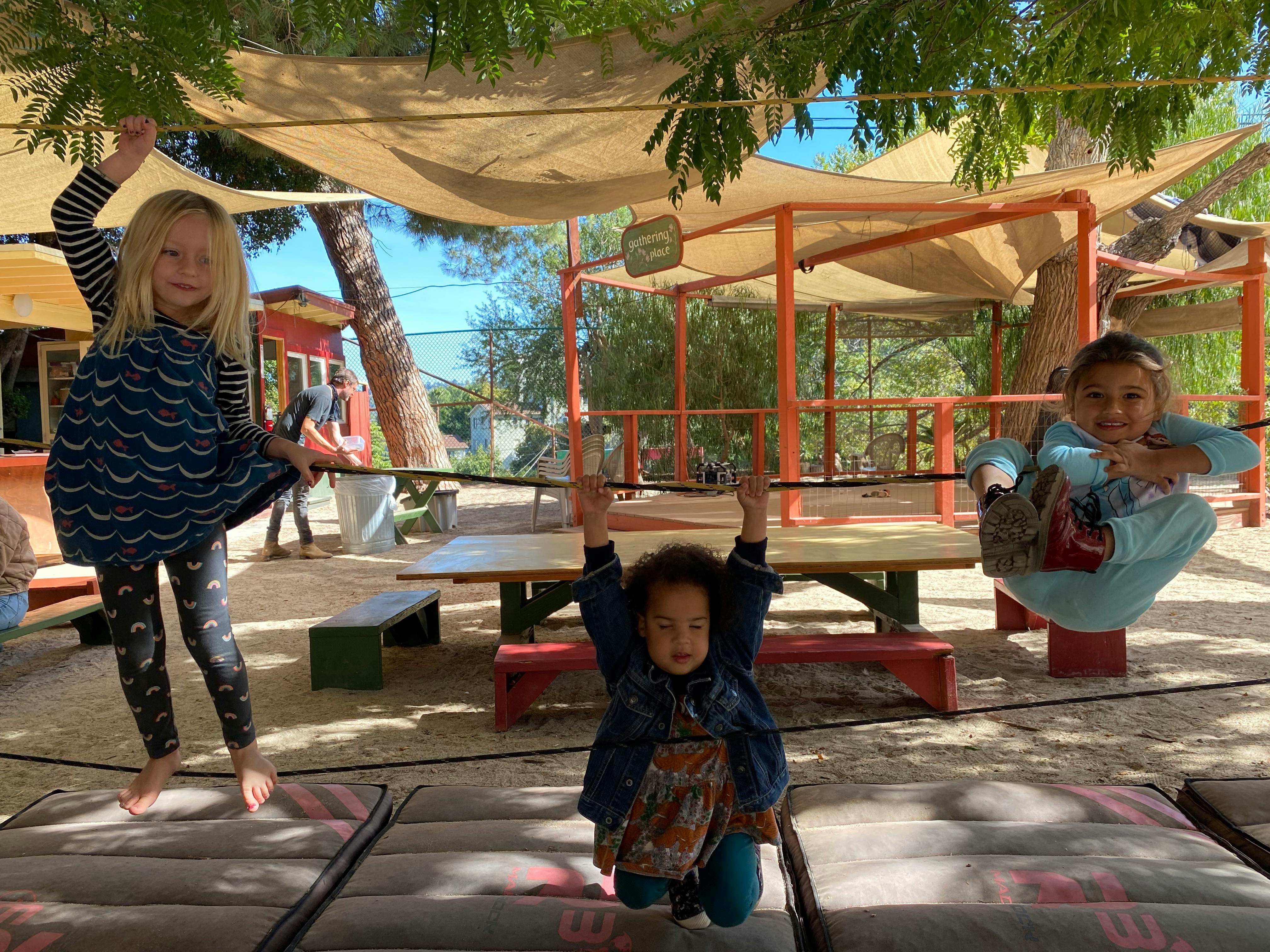 Three children playing outdoors, one on a slackline, one kneeling, one sitting