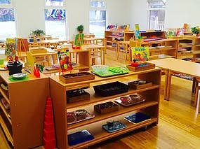 Classroom with wooden tables, chairs, and shelves with learning materials