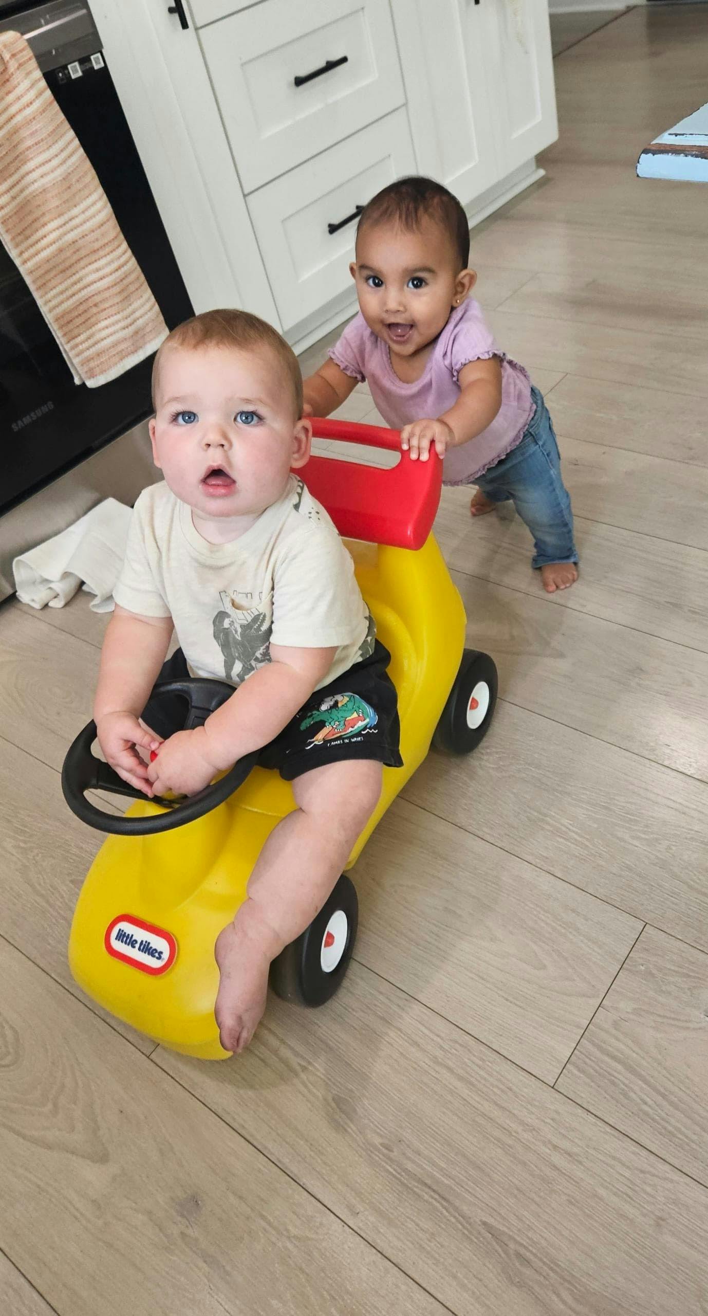 Child sitting in a yellow toy car with another child pushing from behind