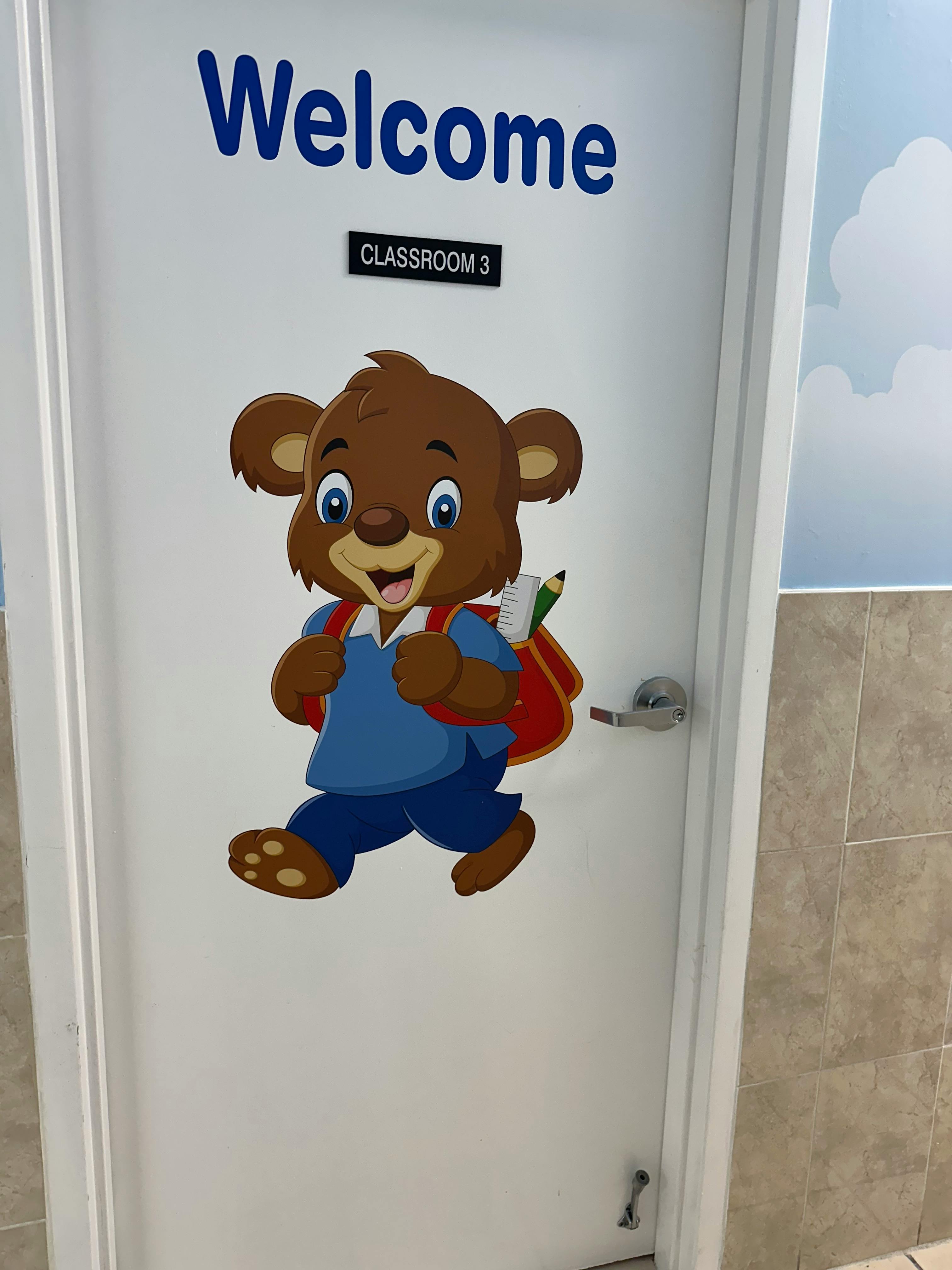 Cartoon bear with backpack on a door that says "Welcome Classroom 3"