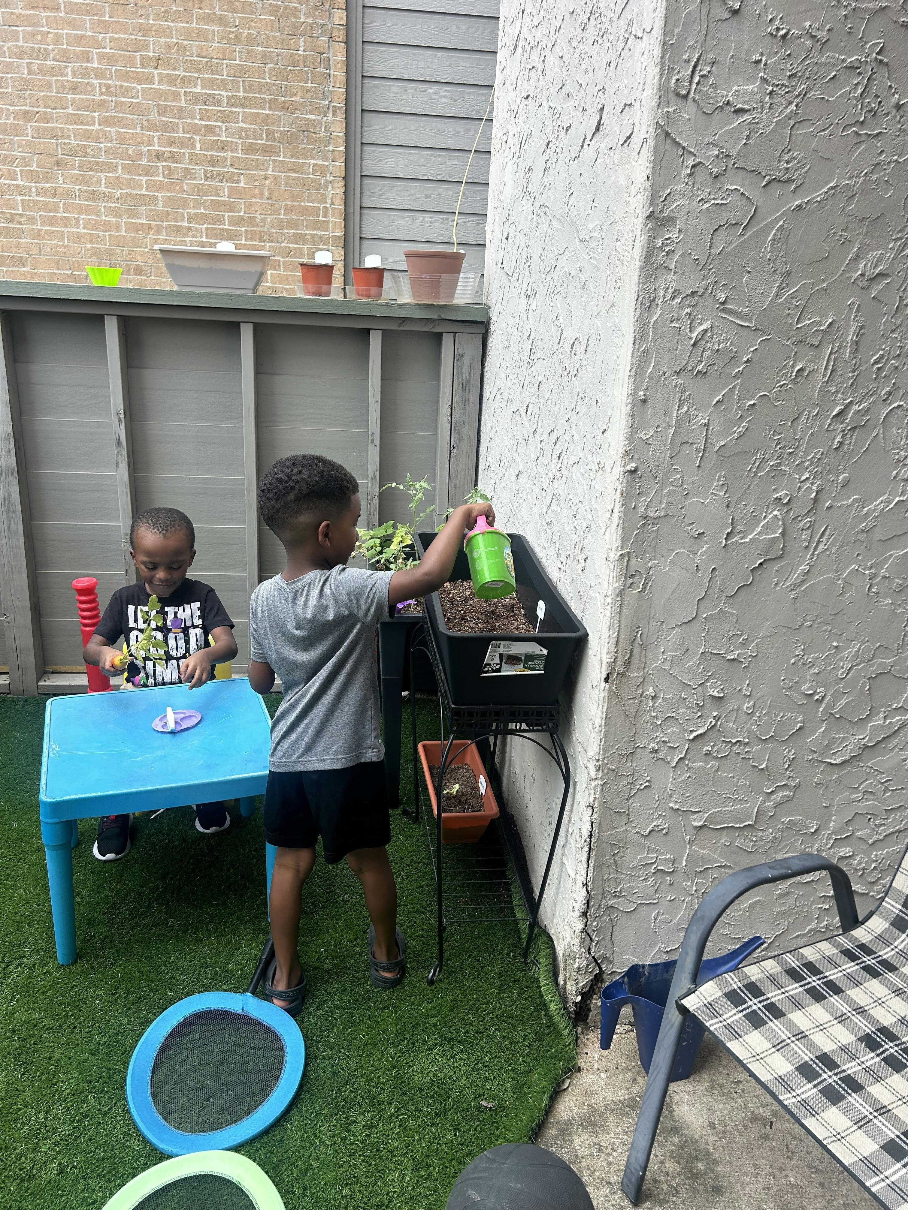 Two children playing outdoors, one watering a plant, one at a table