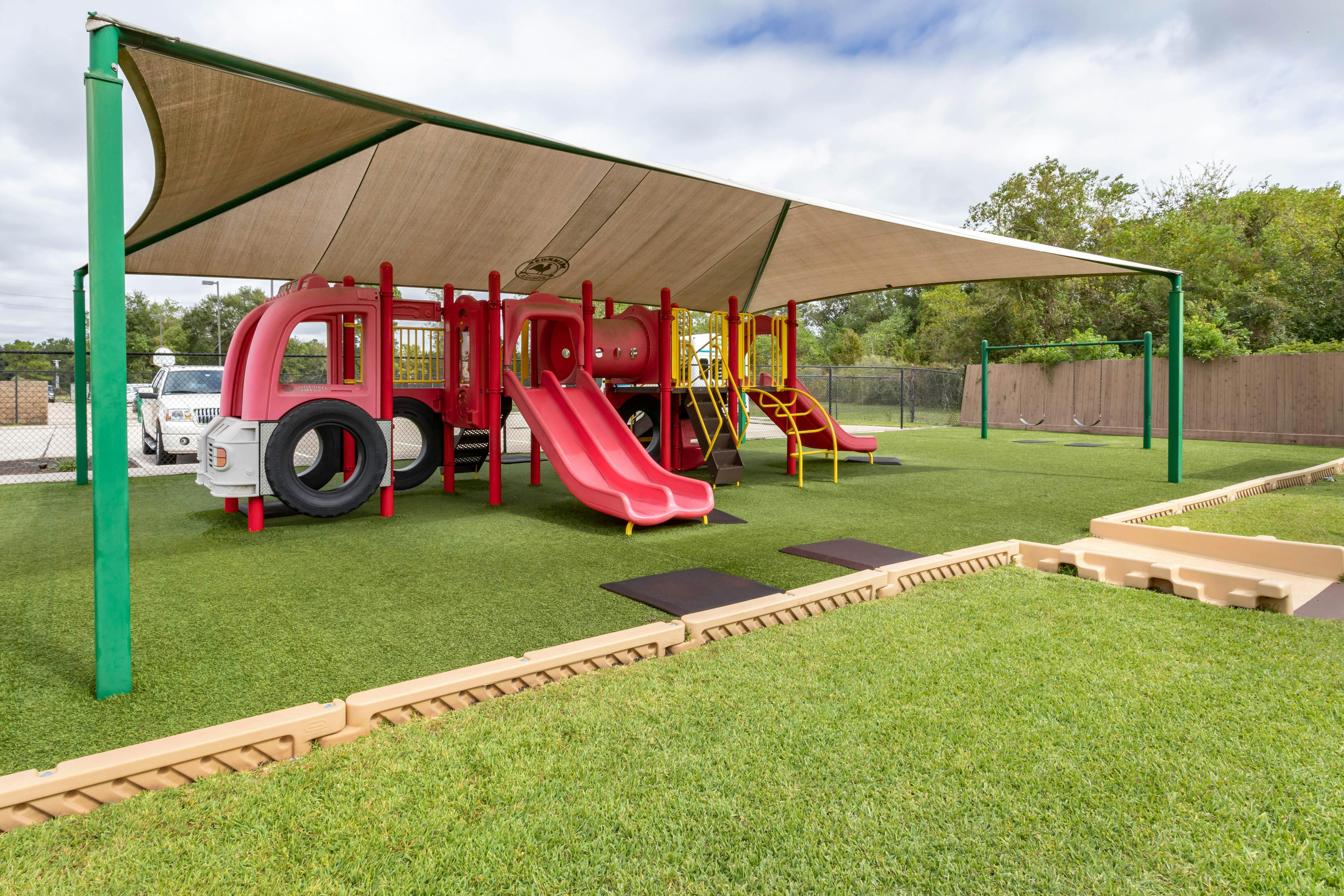 Playground with red fire truck structure, slides, and swings on artificial turf under shade