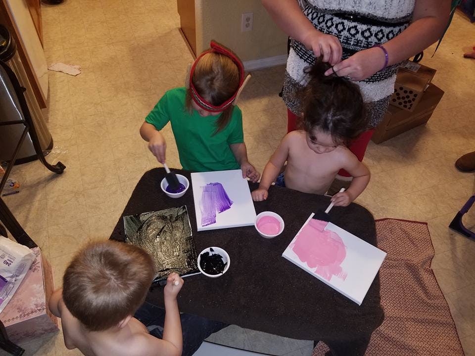 Three children painting at a table with an adult braiding hair