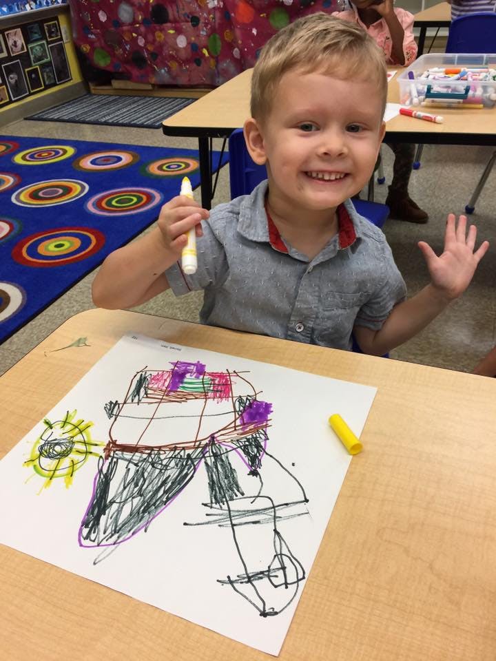Child holding a marker and showing a drawing on paper in a classroom