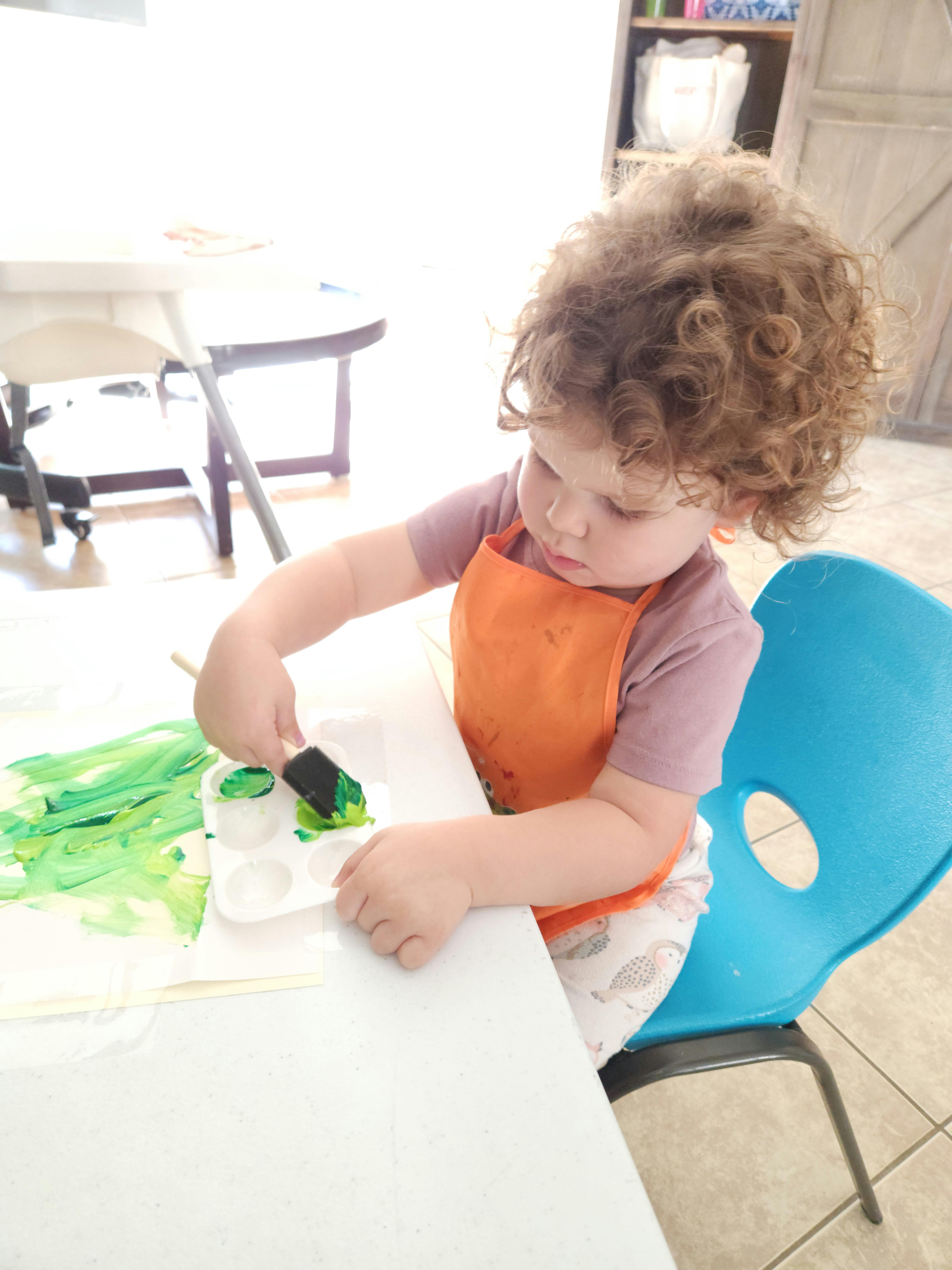 Child in an orange apron painting with green paint at a table