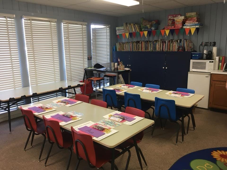Classroom with tables and chairs, books on shelves, and blinds on tables