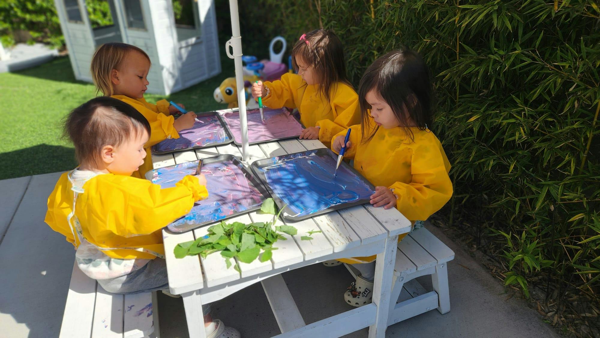 Four children in yellow smocks painting at a picnic table outdoors
