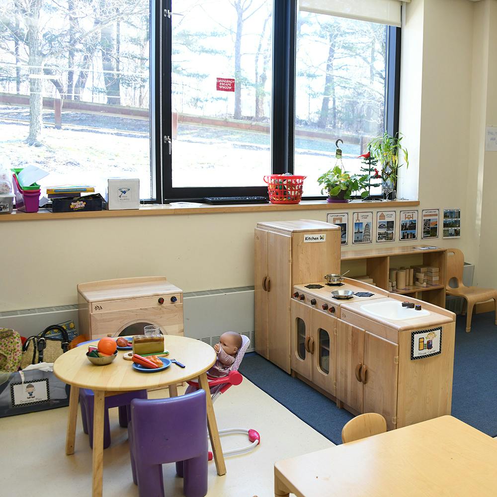 Child in a high chair next to a play table and kitchen set in a classroom