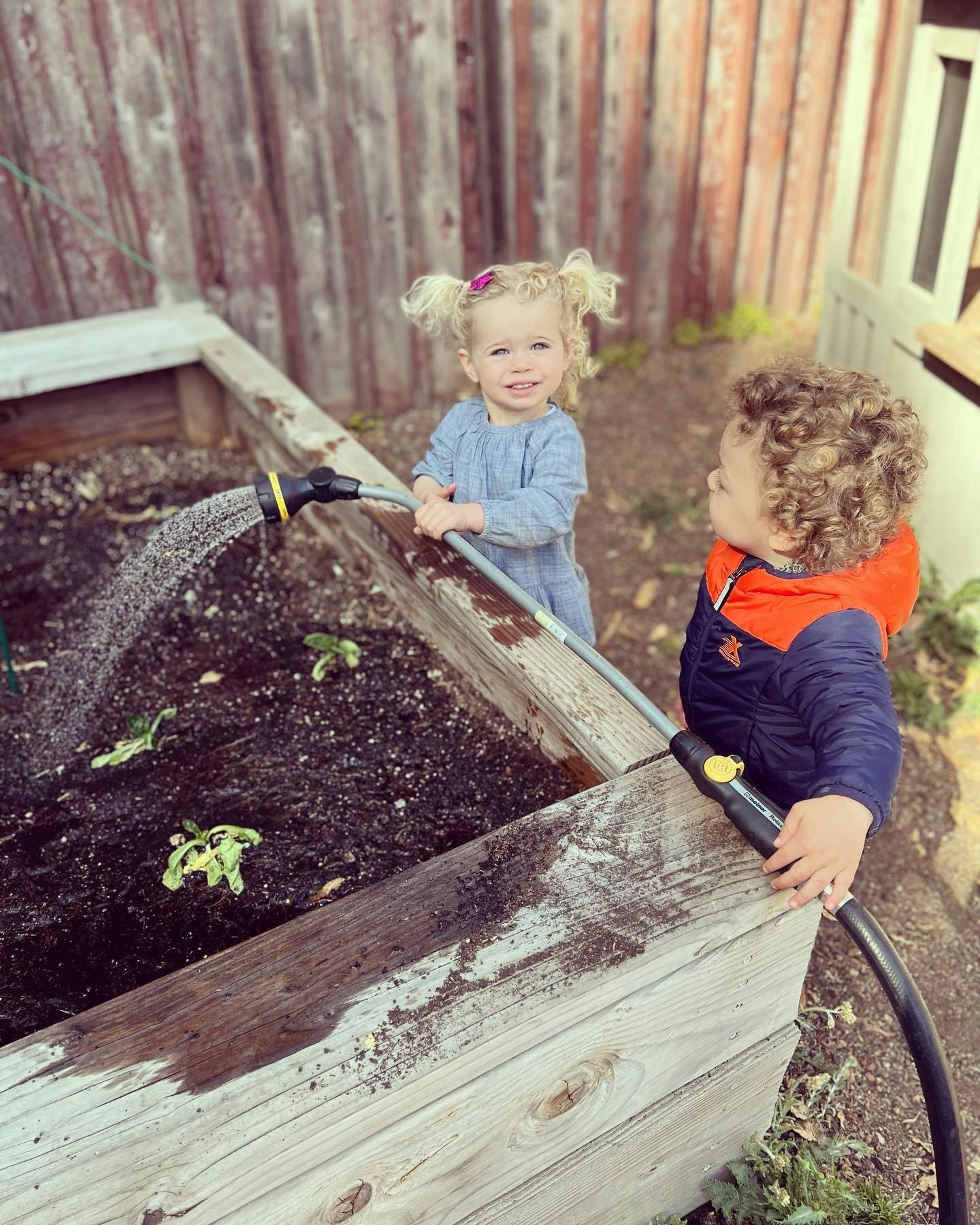 Two children watering plants in a raised garden bed
