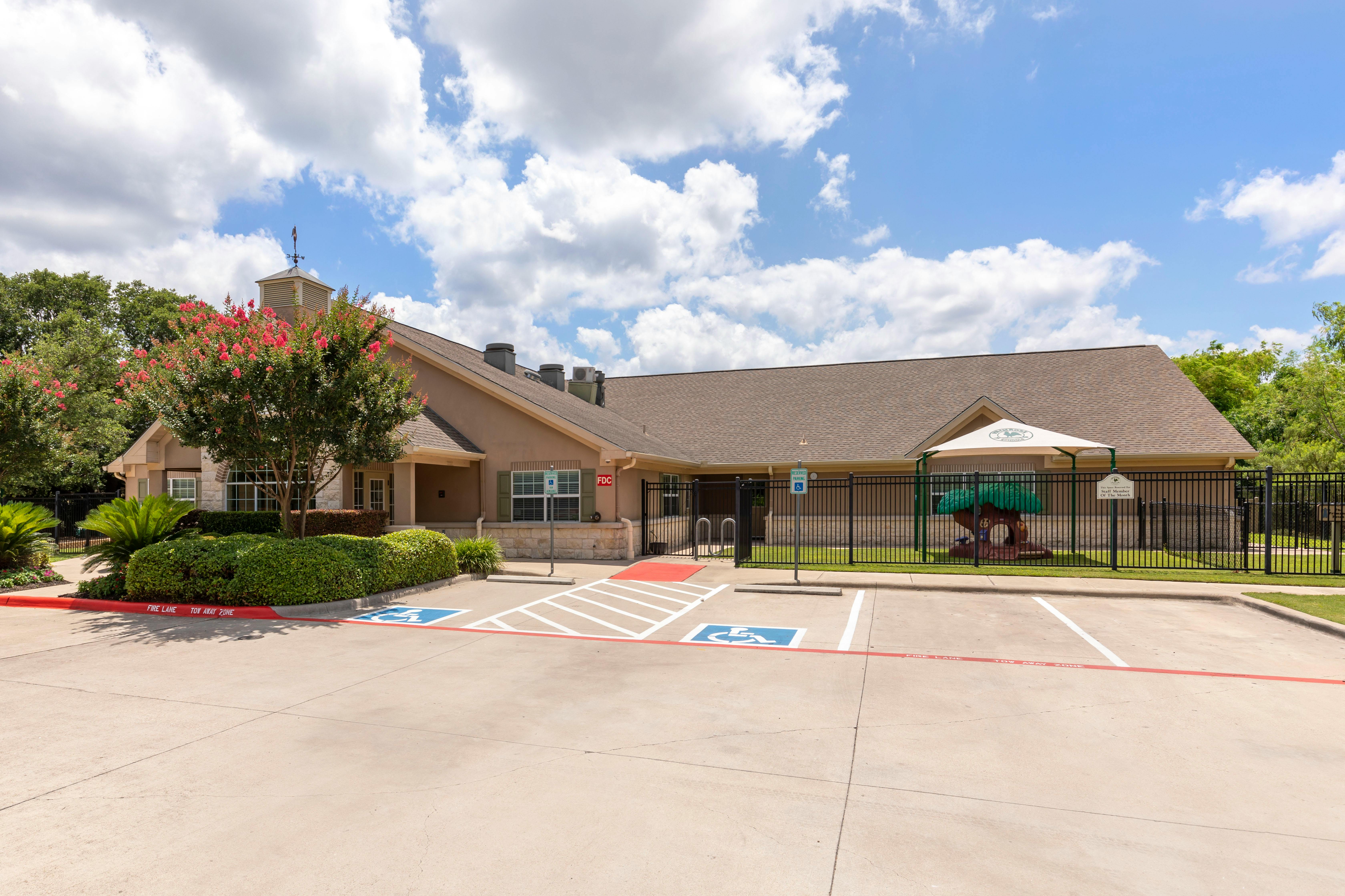 Building exterior with a red flowering tree, parking lot, and fenced playground